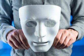 Sindromul impostorului (barbat care tine o masa alba in mana, hanorac gri, fundal albastru)